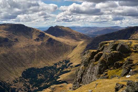 Mountain valley landscape with rocky outcrops, moorland, and distant peaks under dramatic cloudy sky.