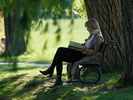 Woman reading a book while seated against a large tree trunk in dappled sunlight.