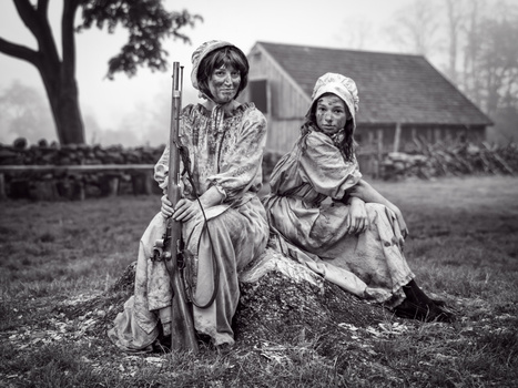 Two figures in period clothing seated outdoors, one holding a rifle, with a rural building and bare tree in background.