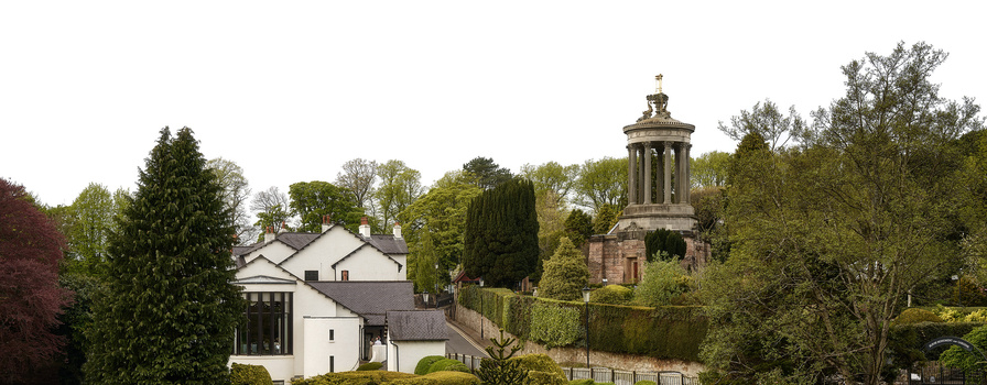 White residential building with classical stone monument tower visible beyond dense green trees and hedges.