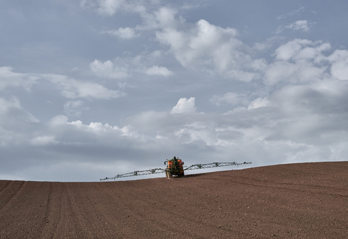 Agricultural sprayer vehicle traversing a brown plowed field under a partly cloudy sky.