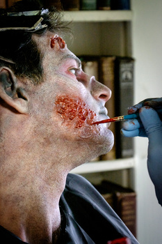 Makeup artist applying red prosthetic wound or special effects makeup to actor's chin and lower face using a brush.