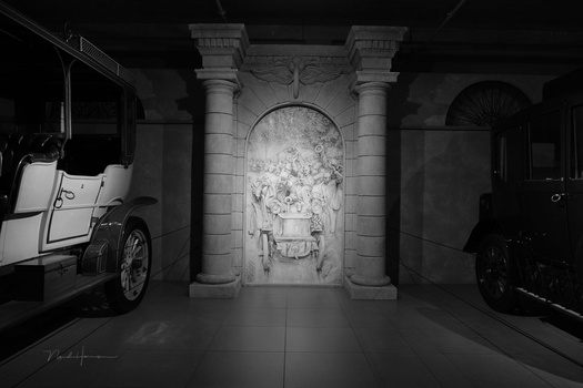 Black and white photograph of a classical stone archway with columns flanked by vintage automobiles in a symmetrical gallery space.