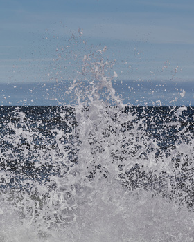 Large ocean wave crashing with white spray and foam against blue water and sky.