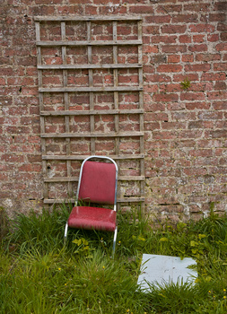 Red metal chair sitting alone on grass against weathered brick wall with empty shelving unit.