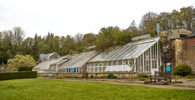 Long Victorian-era greenhouse with glass panels and metal framework extending across manicured lawn.