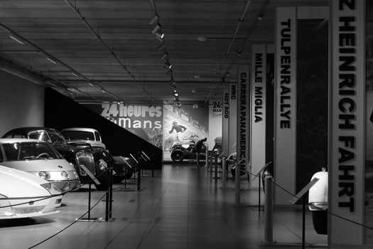 Black and white photograph of a modern automotive museum interior with classic cars on display and vertical text signage on the walls.