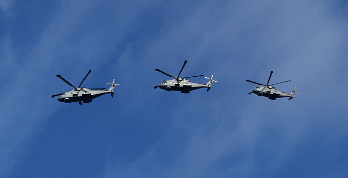Three military transport helicopters in formation flight against a blue sky with white clouds.