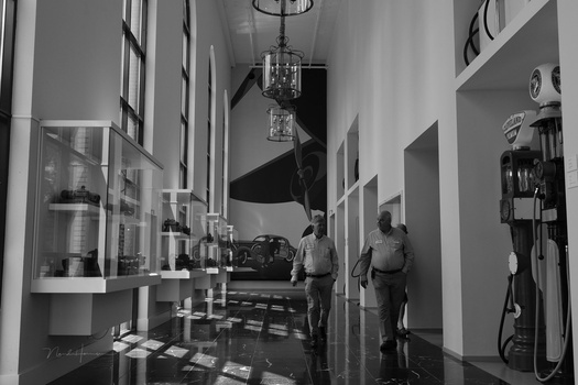 Black and white photograph of a museum or gallery corridor with display cases and dramatic overhead lighting.