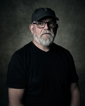 Portrait of a man wearing glasses and a black cap against a textured dark background.