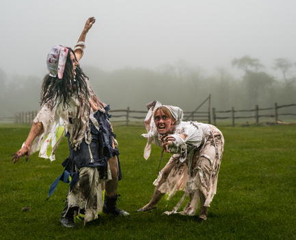 Two people in tattered costumes perform outdoors in a misty field with fencing visible in the background.