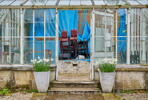Greenhouse entrance with red chairs visible inside, flanked by potted plants with fresh green shoots.
