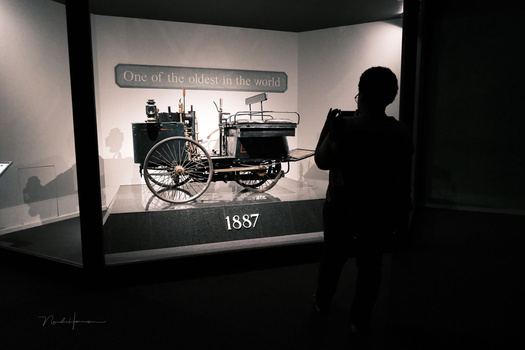 Silhouetted visitor photographing a museum display of an 1887 automobile labeled 'One of the oldest in the world.'
