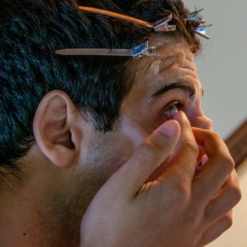 Close-up portrait of a man with glasses examining his face with both hands, shot in warm and cool studio lighting.