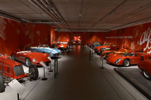 Red sports cars displayed in a museum gallery with bright overhead lighting and Coca-Cola branding on the walls.