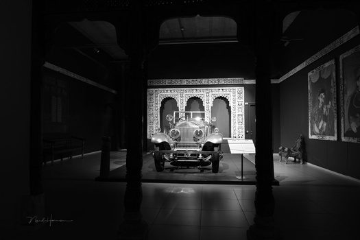Vintage convertible car displayed in a museum gallery, viewed through support columns in dramatic black and white lighting.