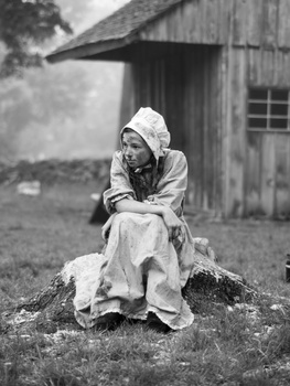 Woman in white headscarf and dress sitting on a tree stump in front of a wooden shed.