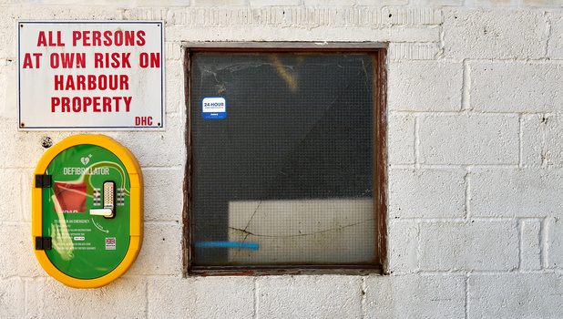White brick wall with red warning sign, green and yellow defibrillator box, and dark window with mesh screen.