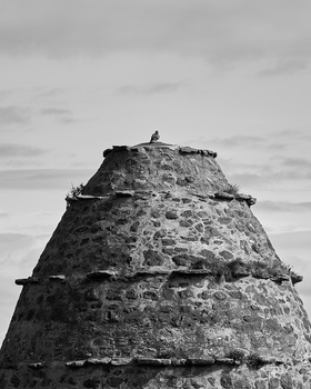 Black and white photograph of a tall, weathered stone tower with a figure standing atop it against a cloudy sky.