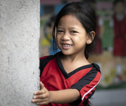 Young girl in red and black sports shirt leaning against white concrete wall, smiling at camera.
