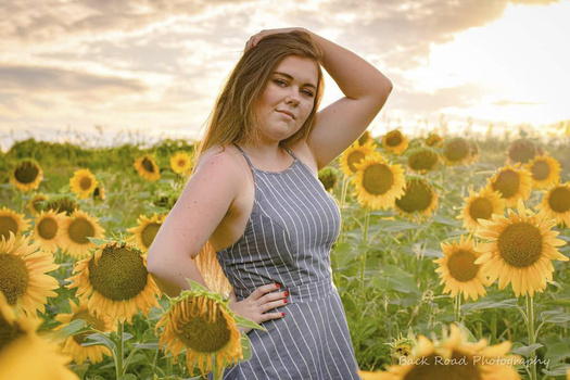 Woman in blue pinstriped dress posing in a sunflower field during golden hour.