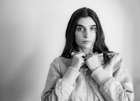 Black and white portrait of a young woman with long dark hair, wearing a light-colored jacket and hands near her collar, gazing directly at camera.