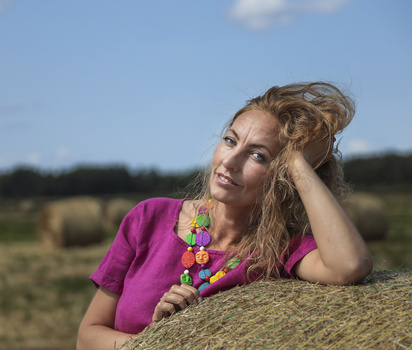 Woman in magenta shirt leaning against hay bale in rural field, wearing colorful beaded necklace.