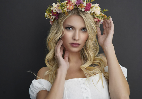 Blonde woman with wavy hair wearing white off-shoulder top and colorful flower crown against dark background.