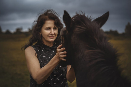Woman with dark wavy hair standing beside a black horse in an open field.
