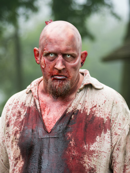 Bearded man with blood on his face and clothing against a blurred outdoor background.