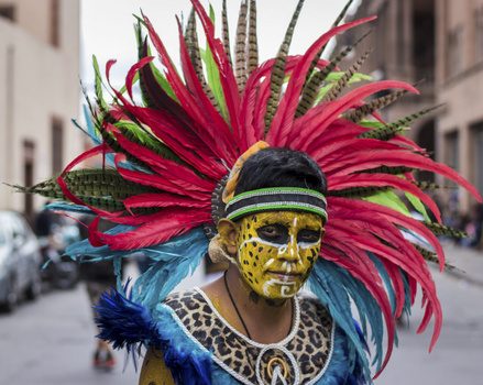 Street performer wearing vibrant red feathered headdress, gold face paint, and ornamental costume against urban background.