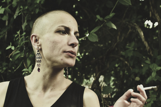Shaved-head person in profile holding a cigarette, framed by lush green foliage.