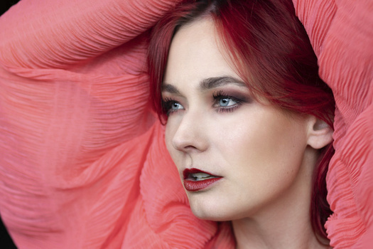 Woman with burgundy hair against pink textured background, wearing dark lipstick and dramatic eye makeup.