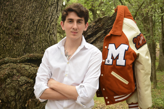 Young man in white shirt posing with rust and cream varsity jacket against tree in forest setting.