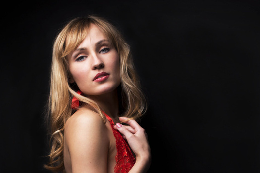 Woman with blonde wavy hair wearing red fabric, posed against dark background with studio lighting.