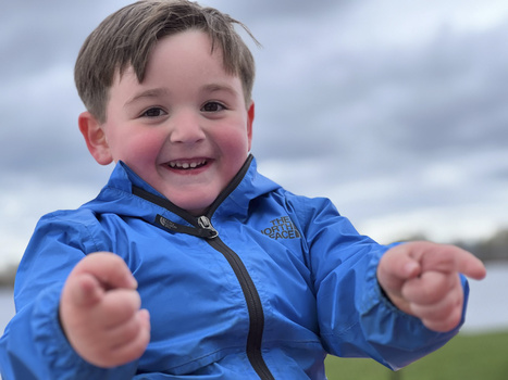 Smiling young child in blue jacket with arms outstretched toward camera against cloudy sky.