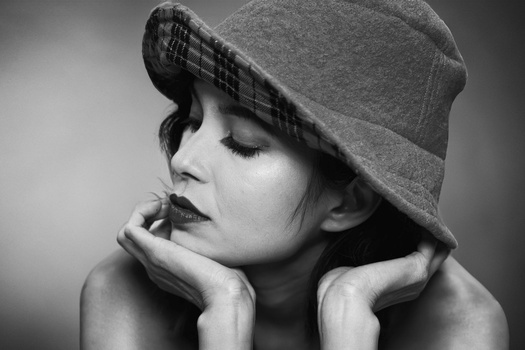 Woman wearing a felt hat with plaid trim, hands framing her face, eyes closed in contemplative expression.
