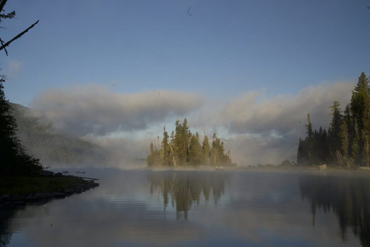 Misty lake at dawn with forested islands and coniferous trees reflected in still water.