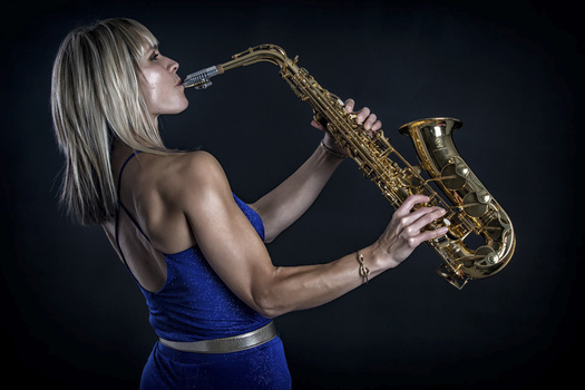 Woman in blue tank top playing saxophone against dark background.