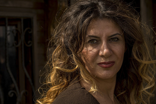 Woman with wavy brown hair lit by warm golden light, looking toward camera with composed expression.