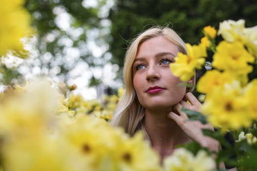 Blonde woman surrounded by yellow flowers, gazing upward with a contemplative expression.
