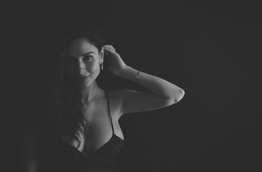 Woman with long dark hair posed against dark background, arm raised to head.
