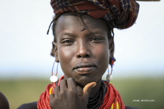 Portrait of a woman wearing red beaded necklaces and a patterned head wrap, with a contemplative expression against a blurred natural background.