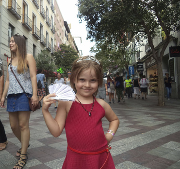 Young girl in red dress posing on a pedestrian street with buildings and trees in background.