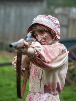 Child in pink patterned clothing aiming a vintage rifle outdoors.