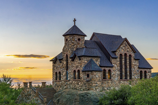 Stone church with round turret and cross at sunset, surrounded by landscaping.