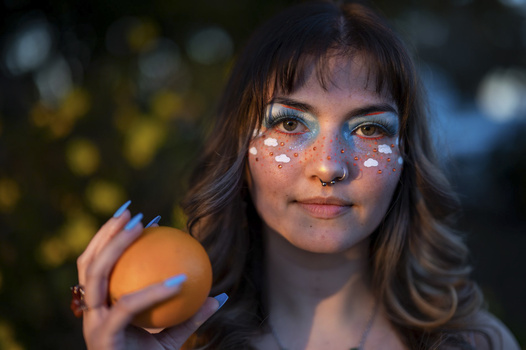 Portrait of a woman with glitter makeup holding an orange against a blurred background.