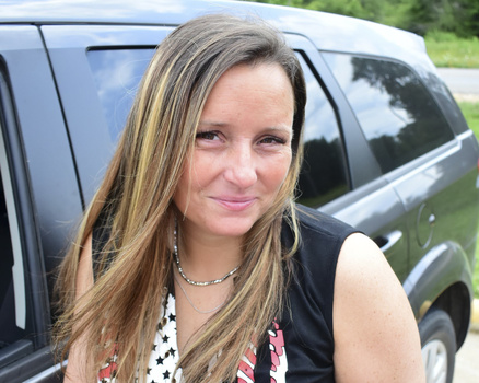 Woman with long brown hair smiling at camera, wearing black sleeveless top and chain necklace, standing beside silver SUV.