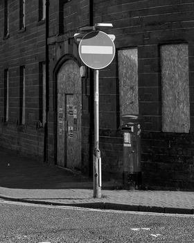Street sign pole casting shadows against weathered brick building facade in black and white.