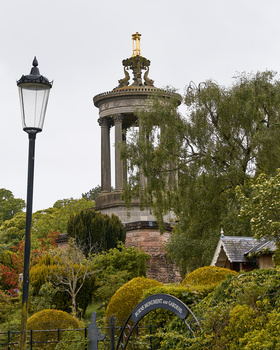 Classical stone rotunda with columned portico rises behind manicured gardens and a vintage street lamp.